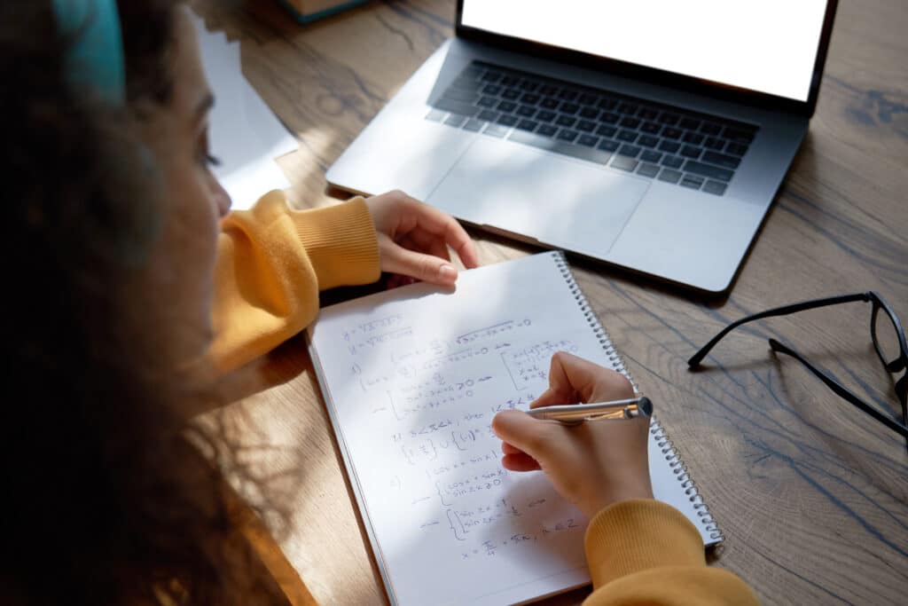 Teen girl college student wear headphones studying from home writing in workbook solving equations learning math sits at desk. Teenage school pupil learn online on laptop, close up over shoulder view.