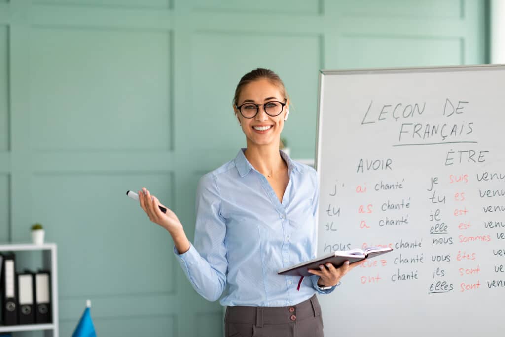 Friendly happy teacher recommending foreign languages school, giving French lesson, standing near whiteboard and smiling at camera. Positive young tutor satisfied with educational services