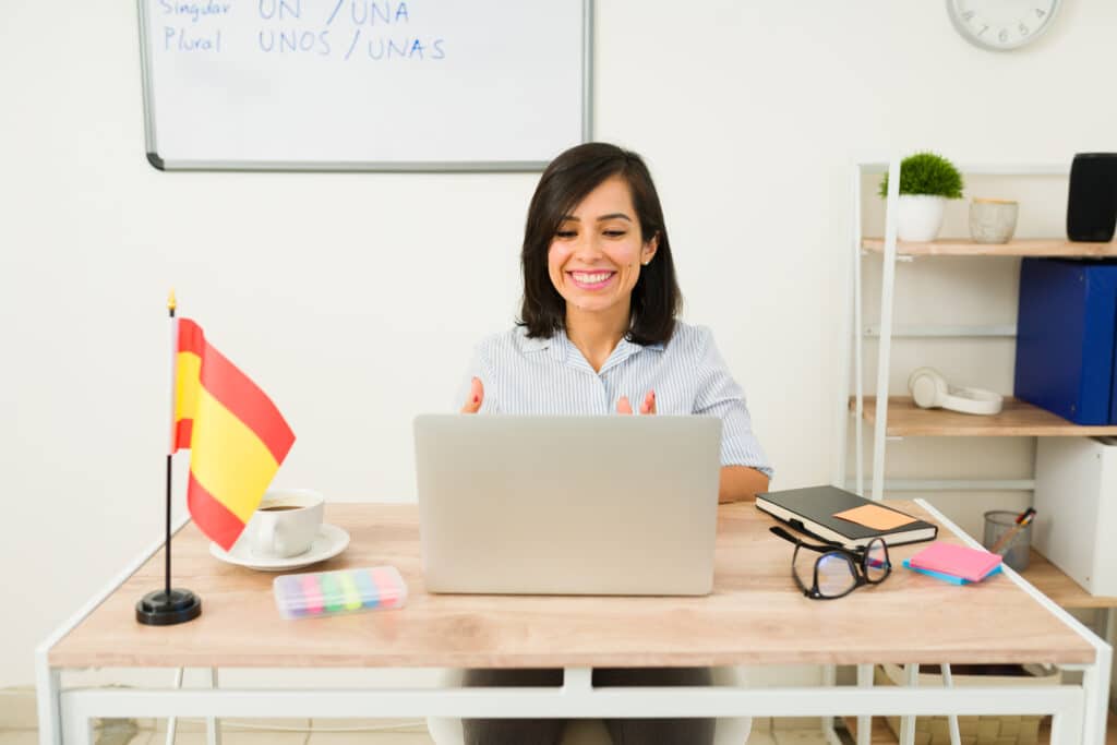Smiling beautiful woman teacher working on the laptop sitting at her classroom desk and looking happy teaching Spanish lessons